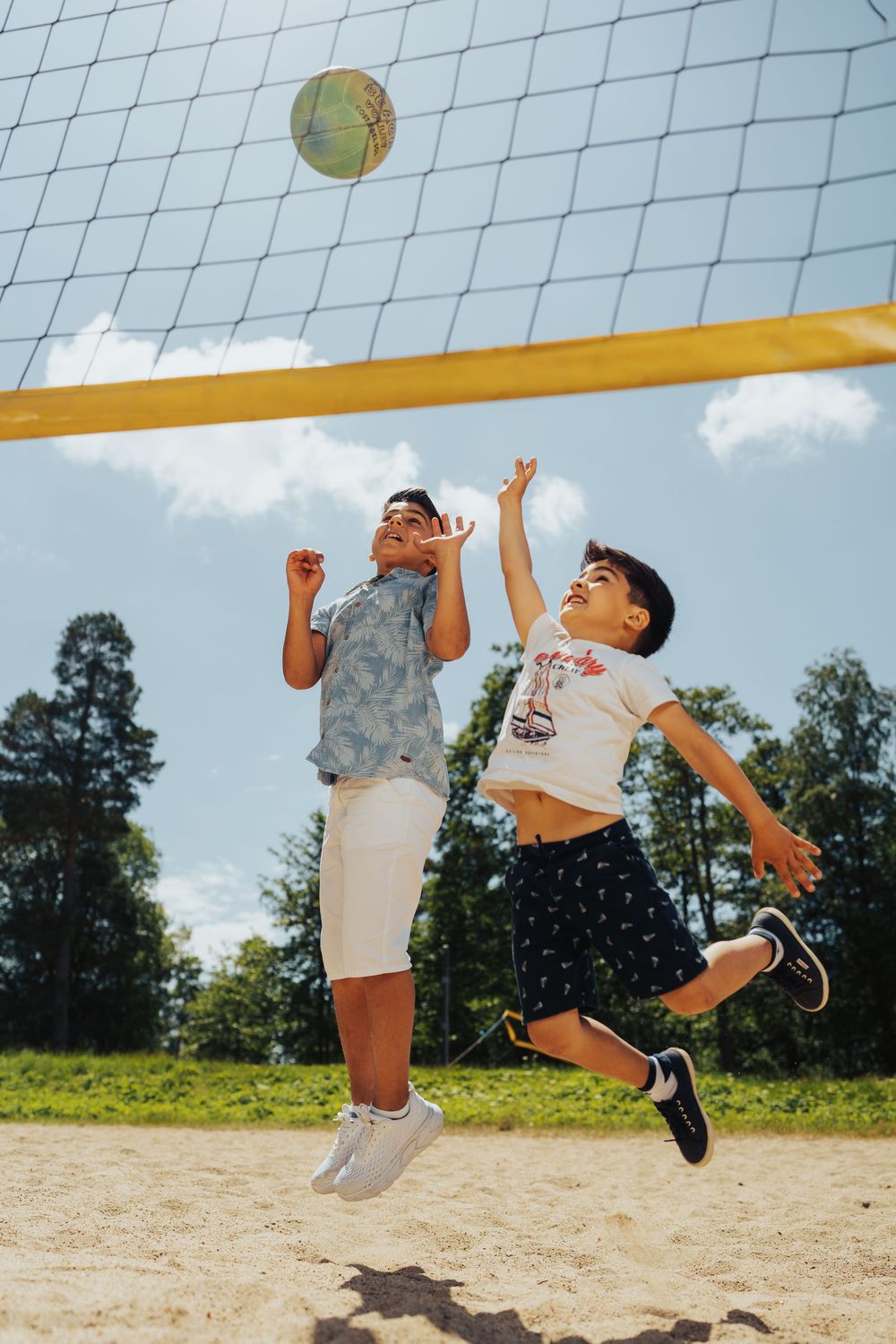 Child jumping to hit volleyball over net on sandy beach court during summer day.