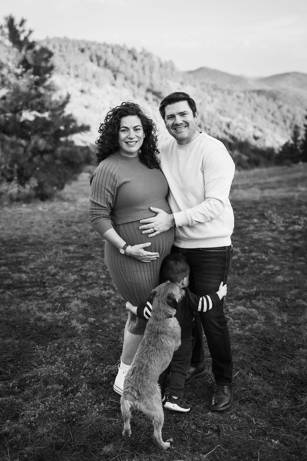 A family smiles in maternity photograph, child and dog joining in family hug in Denver foothills