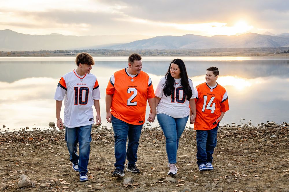 Family of four walking together wearing football jerseys by lakeside at sunset.