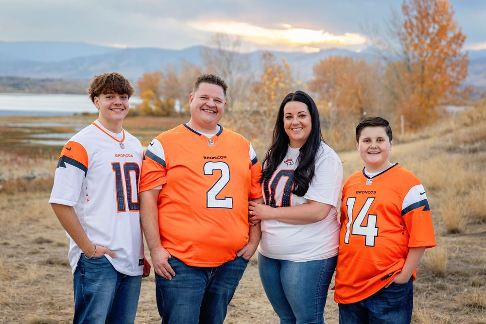 A group wearing Denver Broncos jerseys poses together against an autumn lakeside backdrop.