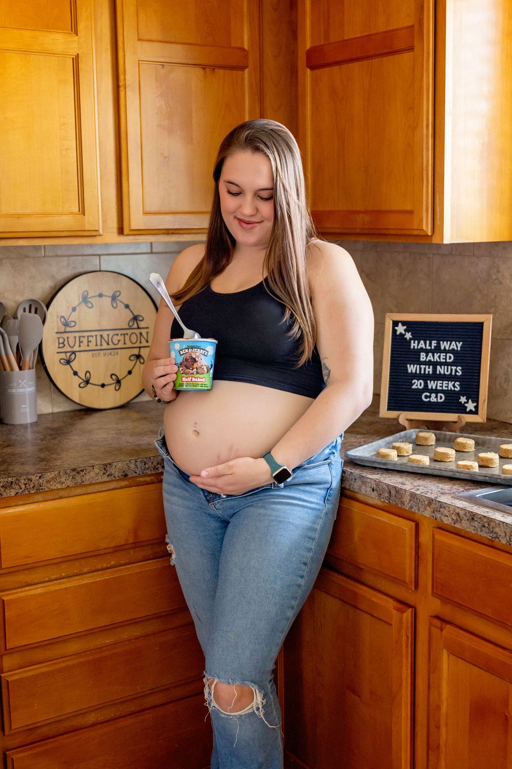 Expectant mother holding ice cream poses in kitchen with wooden cabinets and decorative sign.