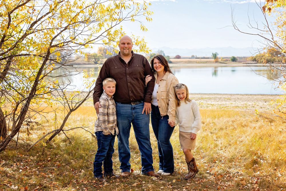 Family poses together in front of autumn trees and lake during golden hour.