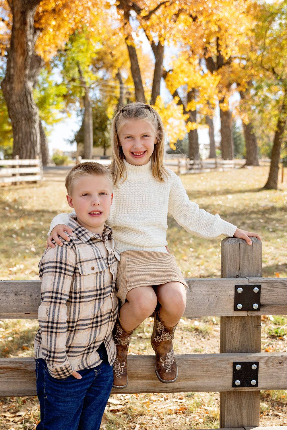 Two children pose on wooden fence during autumn photoshoot with golden leaves in background.