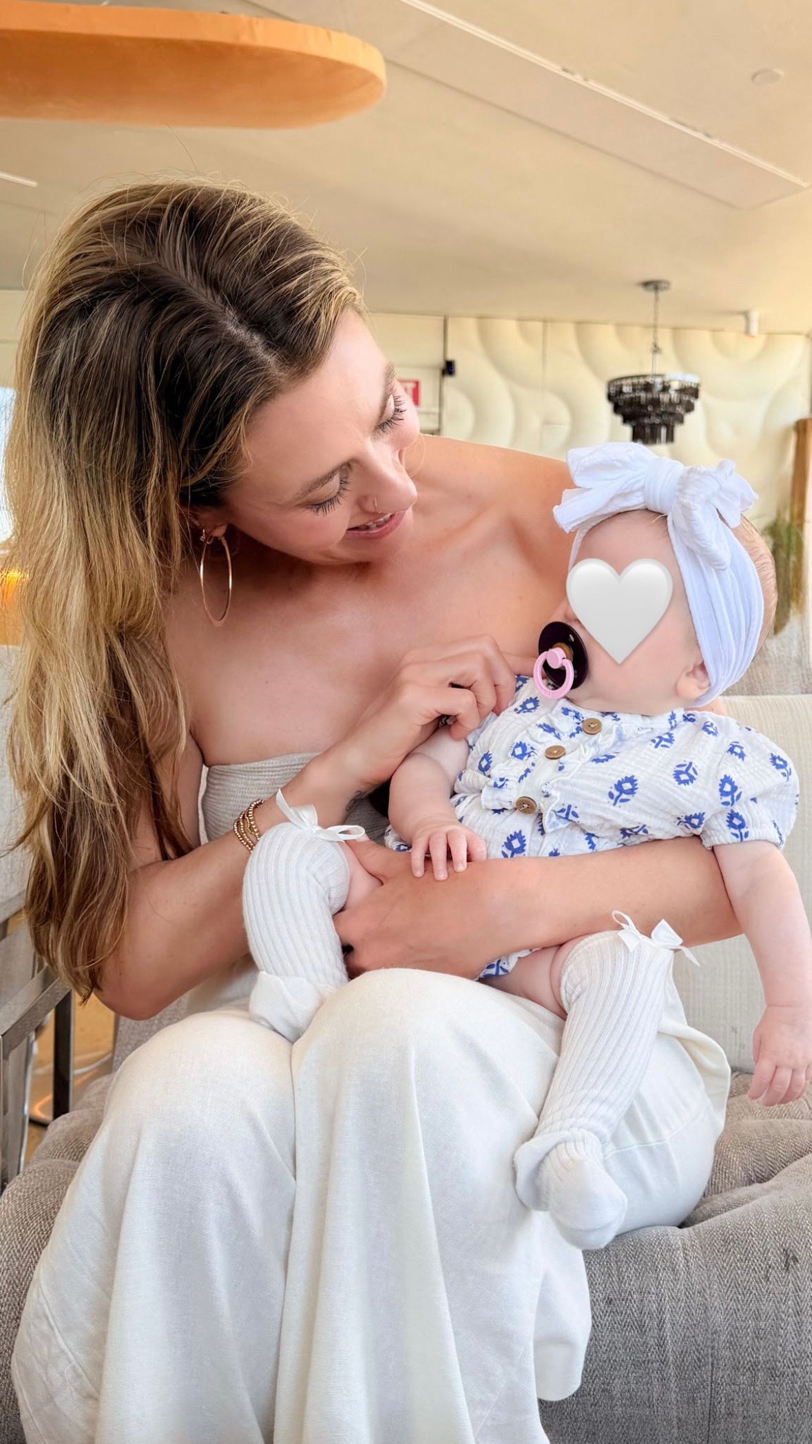 A young woman in white outfit holds a baby wearing a floral onesie and white bow headband with a pacifier.