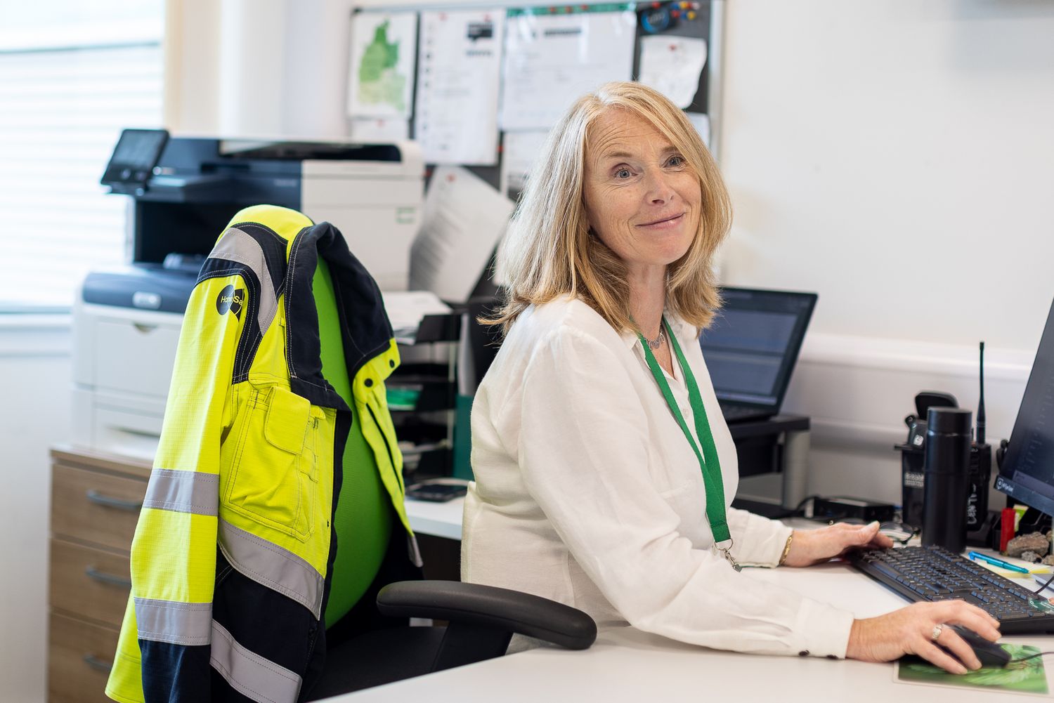 Office worker in high visibility safety vest working at desk with computer monitors and paperwork.