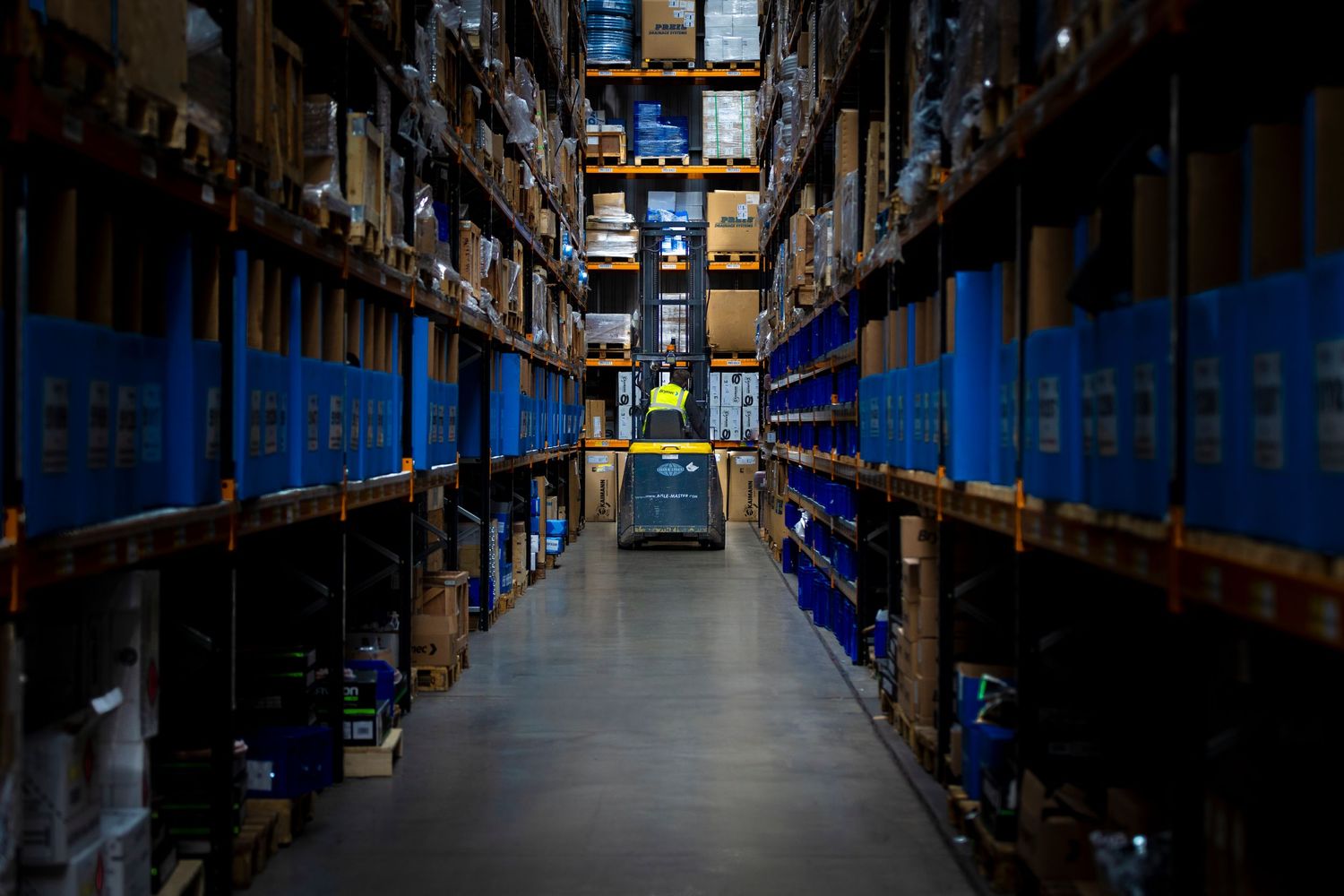 Long warehouse aisle with tall storage racks and blue shelving units illuminated by overhead lighting.
