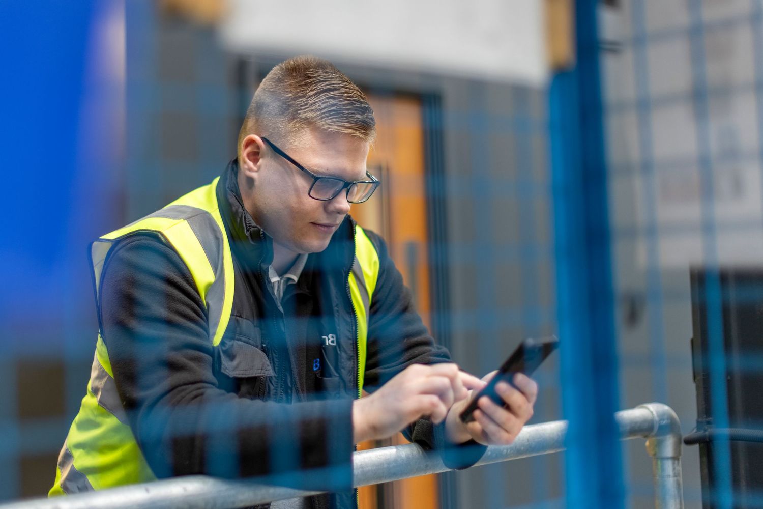 Worker in safety vest using mobile phone while standing in industrial setting.