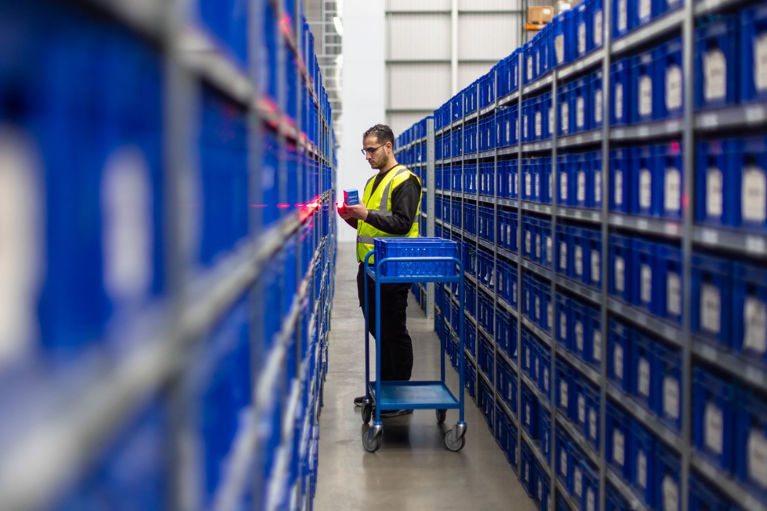 Worker in high-visibility vest organizing inventory between blue storage shelves in warehouse facility.