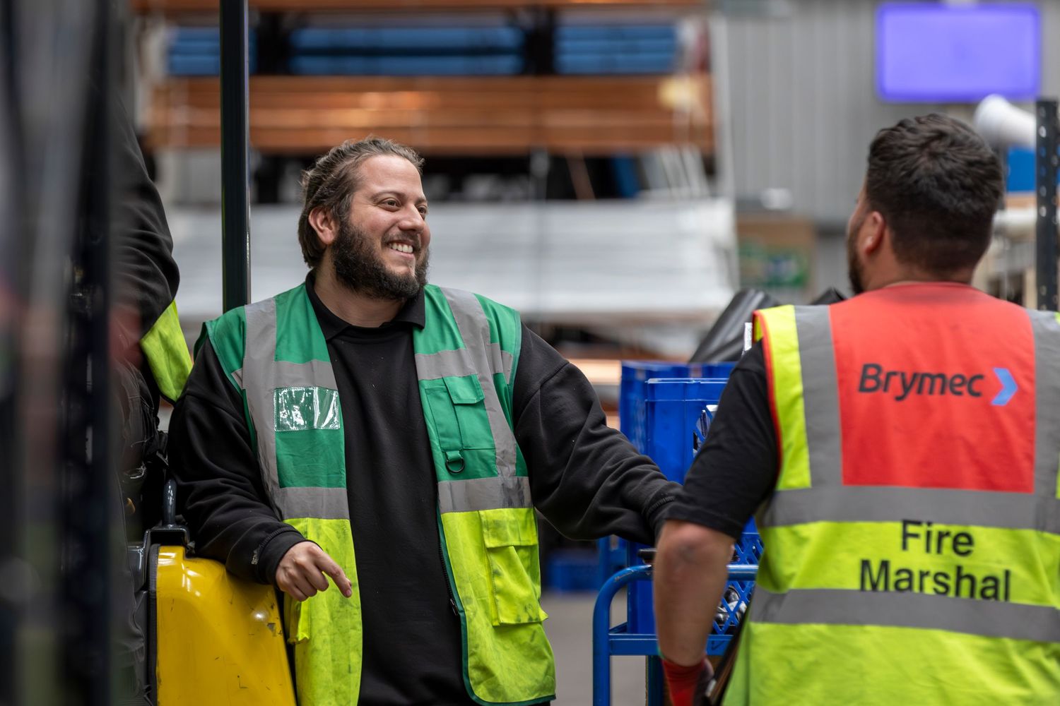 Two warehouse workers in high visibility safety vests having a conversation in industrial setting.