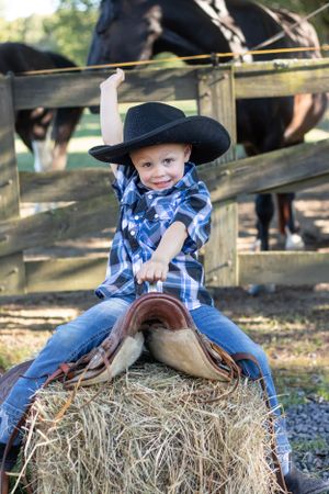 Young cowboy in blue plaid shirt and hat sitting on hay bale near horse fence.