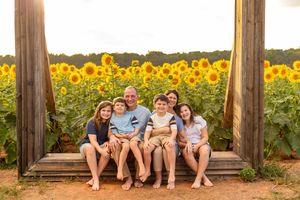 Family of six sitting on a wooden bench surrounded by vibrant sunflower fields in golden hour light.