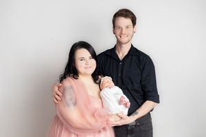 A couple in a professional newborn photo session, woman in pink gown and man in black shirt holding their swaddled baby.