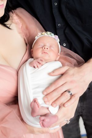 Newborn baby girl swaddled in white with floral headband, cradled by parents in pink dress and black shirt.