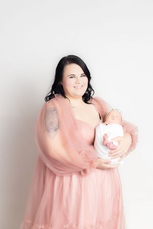 Smiling mother in pink tulle dress holding sleeping newborn baby against white background.