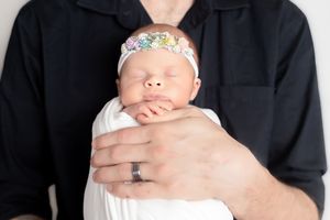 A father in a black shirt cradles his sleeping newborn baby girl wrapped in white with a floral headband.