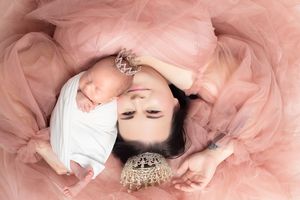 Mother and newborn baby lying together on pink tulle fabric, both wearing crowns in an elegant portrait.