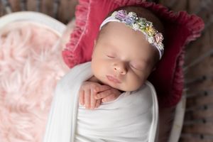 Sleeping newborn baby girl wrapped in white swaddle with floral headband in pink basket.