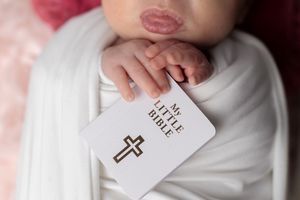 Newborn baby wrapped in white swaddle holding a small My Little Bible book with a cross, with pink kiss marks on forehead.