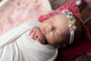 Sleeping newborn baby girl swaddled in white wrap wearing floral headband on pink fur backdrop.