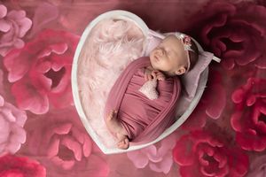 Newborn baby girl swaddled in pink wrap sleeping in heart-shaped bowl with floral pink background.