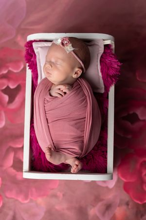 Newborn baby girl swaddled in pink wrap sleeping in a tiny white bed surrounded by pink floral backdrop.
