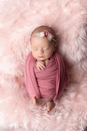 Sleeping newborn baby girl swaddled in pink wrap with floral headband on soft pink fur backdrop.