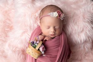 Sleeping newborn baby girl wrapped in pink, wearing floral headband, holding small basket on fluffy pink fur backdrop.