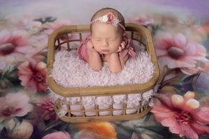 Sleeping newborn baby girl in a wicker basket with pink fluffy blanket and floral headband on a flower backdrop.