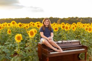 A smiling girl sits barefoot on a brown piano surrounded by vibrant yellow sunflowers in a field at golden hour.