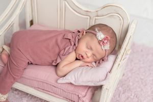 Newborn baby girl sleeping peacefully in pink wrap on miniature white bed with floral headband.