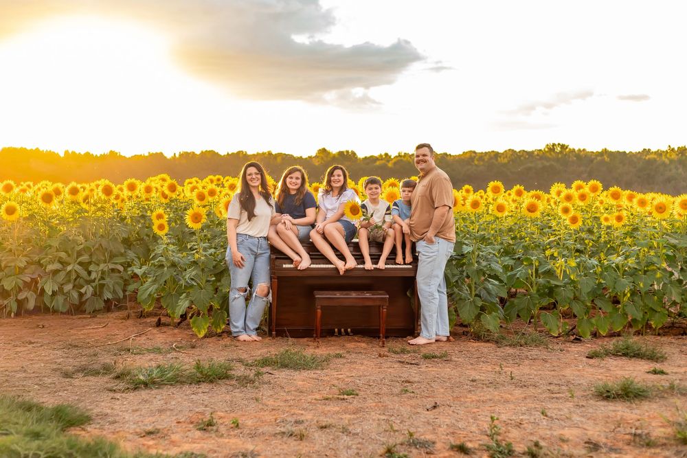 Family of six poses around a vintage piano in a golden sunflower field at sunset.