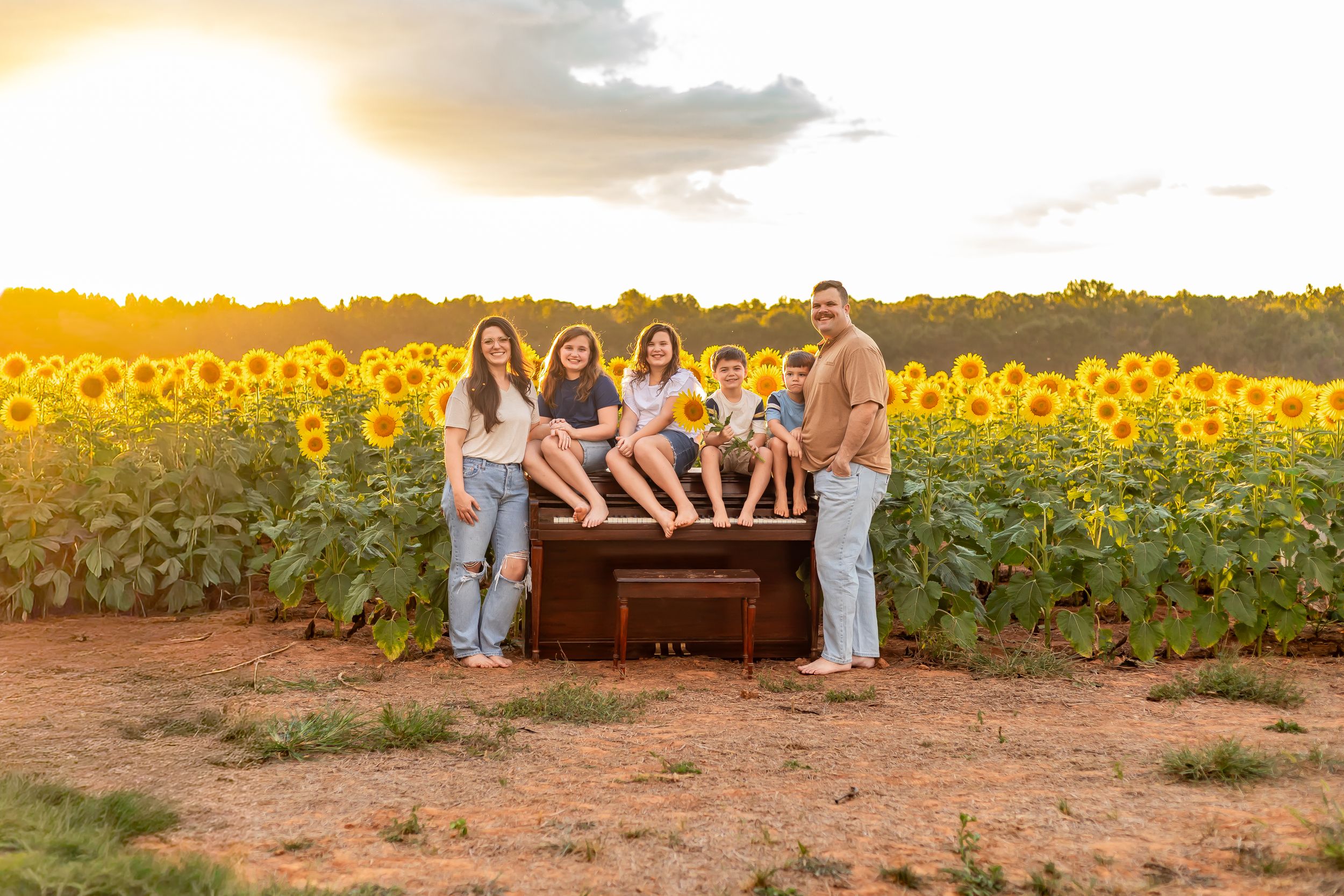 Family of six poses around a vintage piano in a golden sunflower field at sunset.