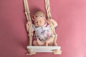 Sleeping newborn baby in floral onesie resting on a white macrame swing against a pink background.