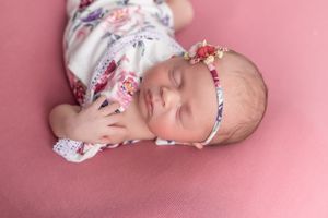 Sleeping newborn baby girl in floral outfit and pink headband lying on pink backdrop.