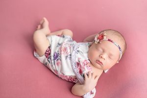 Sleeping newborn baby girl in floral romper and headband on pink backdrop.