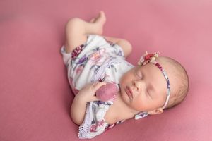 Sleeping newborn baby girl in floral romper and pink headband on pink backdrop.