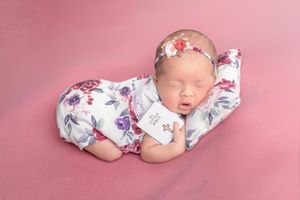 Sleeping newborn baby wrapped in floral swaddle with matching headband on pink background.