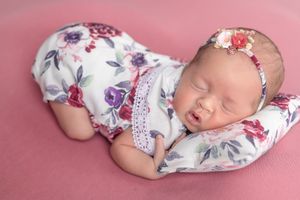 Sleeping newborn baby in floral romper and headband on pink backdrop.