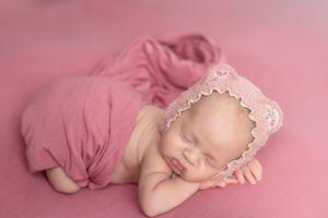 Sleeping newborn baby girl in pink lace bonnet wrapped in soft pink fabric on pink background.