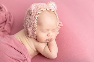 Sleeping newborn baby wearing a pink lace bonnet, posed on a soft pink blanket in a serene studio setting.