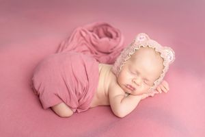 Sleeping newborn baby wrapped in pink cloth wearing a pink bear bonnet on a soft pink background.
