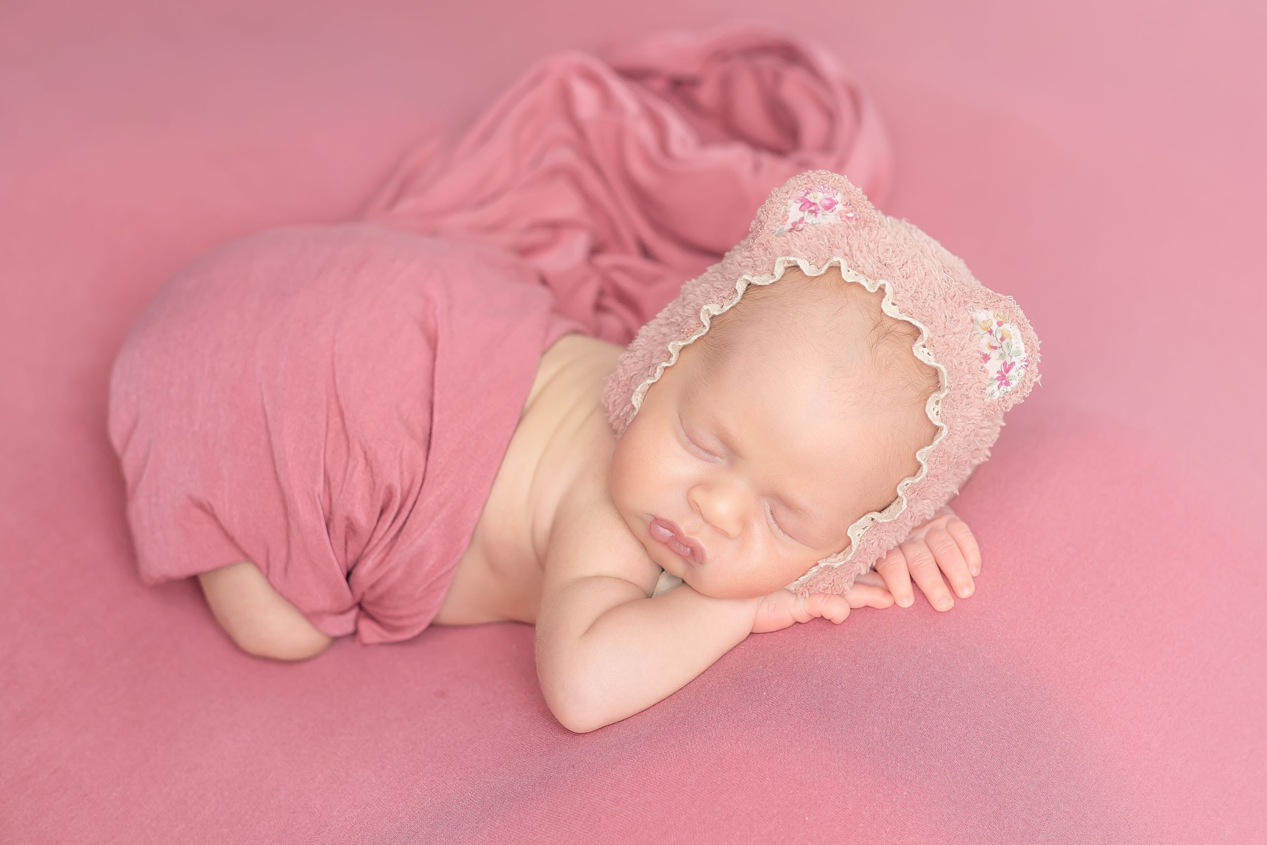 Sleeping newborn baby wrapped in pink fabric wearing a lace bear bonnet on a pink background.