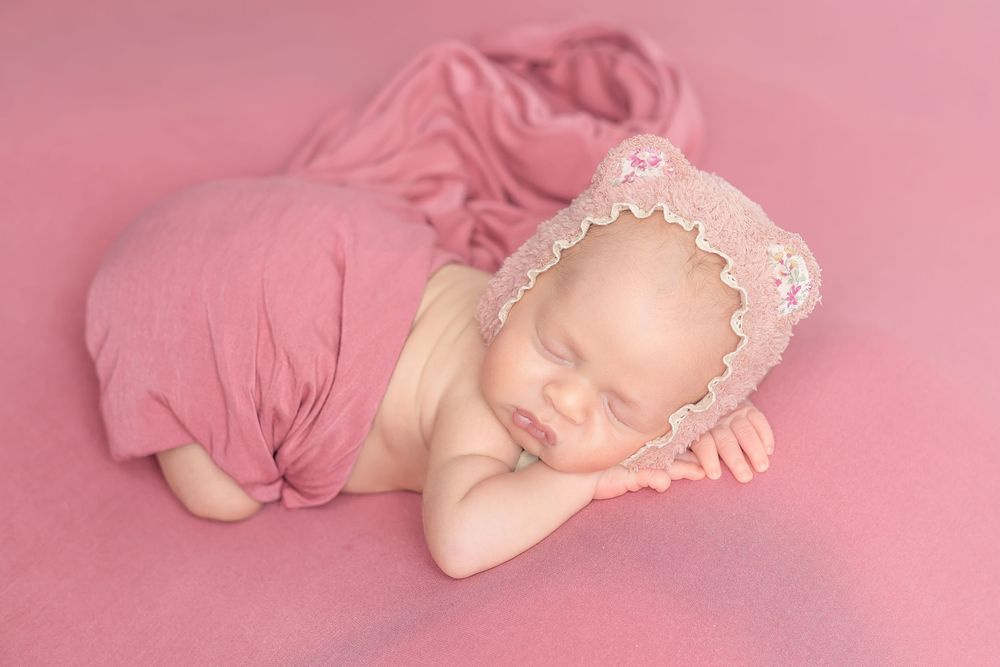Sleeping newborn baby wrapped in pink fabric wearing a lace bear bonnet on a pink background.