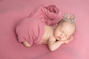 Sleeping newborn baby wrapped in pink fabric wearing a tiny silver crown on a pink background.