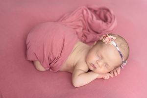 Sleeping newborn baby girl wrapped in pink fabric with floral headband on a pink background.