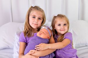 Three siblings in purple dresses share a tender moment with a newborn.
