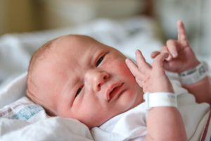 Newborn baby in white clothing with hospital bracelet making expressive face.