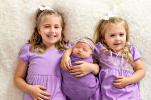 Three siblings in matching purple dresses pose with their newborn sibling.