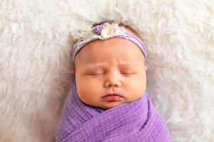 Sleeping newborn wrapped in purple swaddle with floral headband on white furry backdrop.
