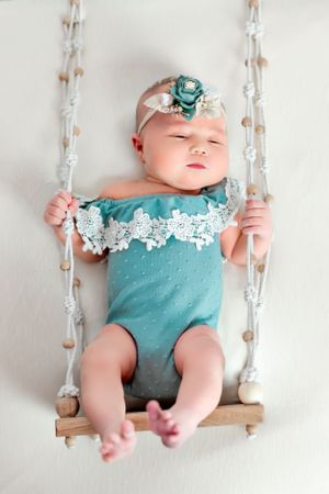 Sleeping baby in teal lace outfit resting on small wooden swing.